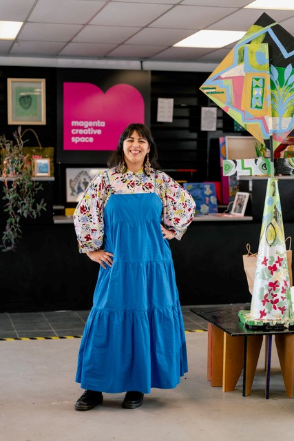 Alicia Rocha Treviño is wearing a long blue dress and standing in front of the pink and white Magenta Creative Spaces logo